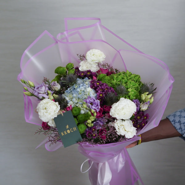 Bouquet of flowers with a pink wrap held by a person, against a neutral background.
